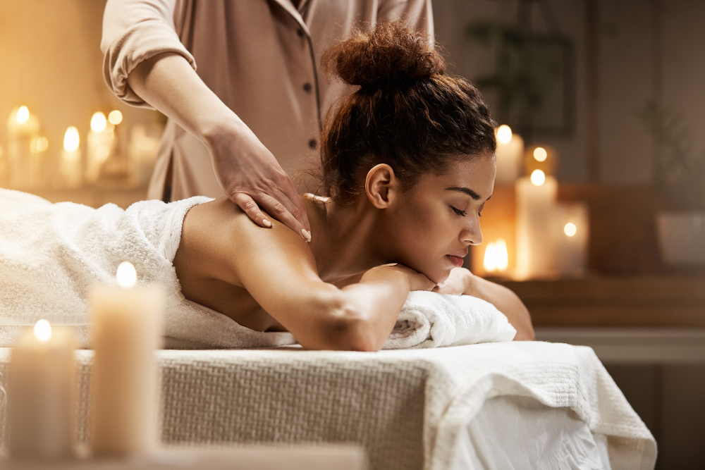 Uma mulher africana sorrindo de olhos fechados, aproveitando uma massagem relaxante em um spa, deitada em uma maca com uma expressão serena e tranquila.