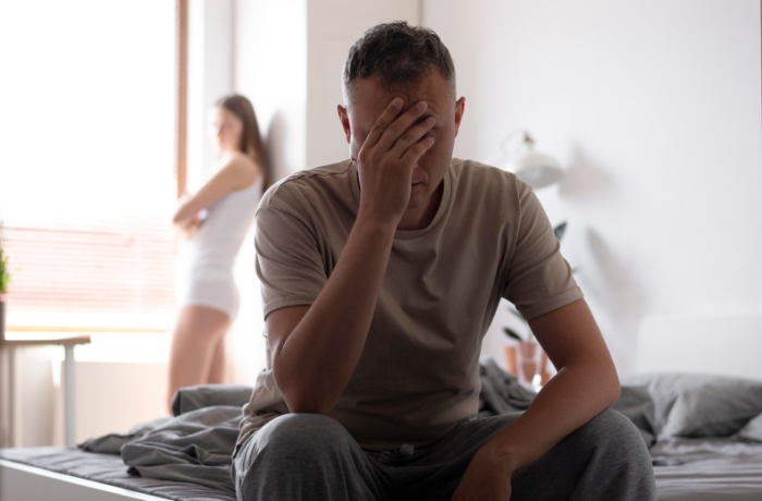 Homem sentado na cama com expressão de frustração, representando ansiedade de desempenho e dificuldades emocionais em relacionamentos íntimos.