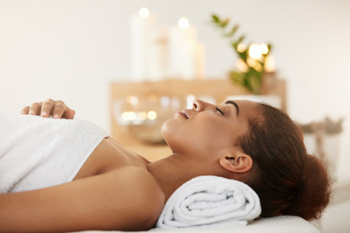 Mulher deitada em uma maca de massagem com toalha branca, em ambiente relaxante com velas acesas e decoração suave ao fundo.